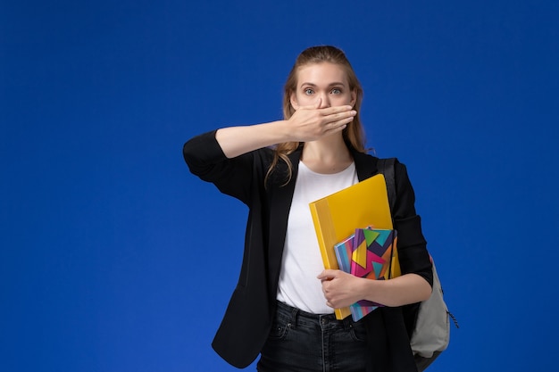 Studentessa di vista frontale in giacca nera che indossa lo zaino che tiene i file con i quaderni sulla lezione dell'università universitaria della parete blu chiaro