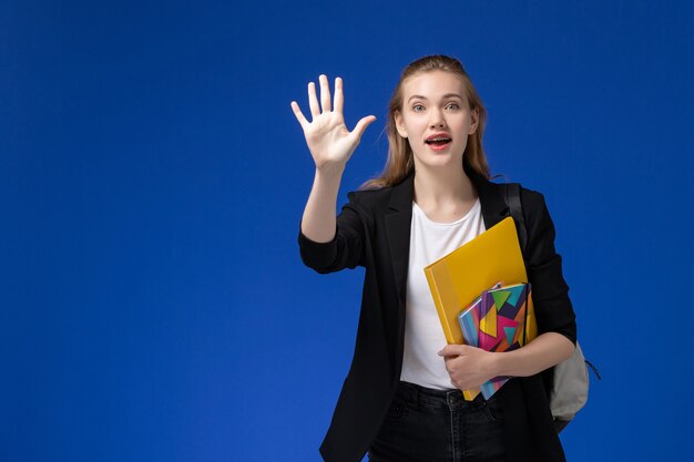 Studentessa di vista frontale in giacca nera che indossa lo zaino che tiene i file con i quaderni sulla lezione dell'università universitaria della parete blu chiaro