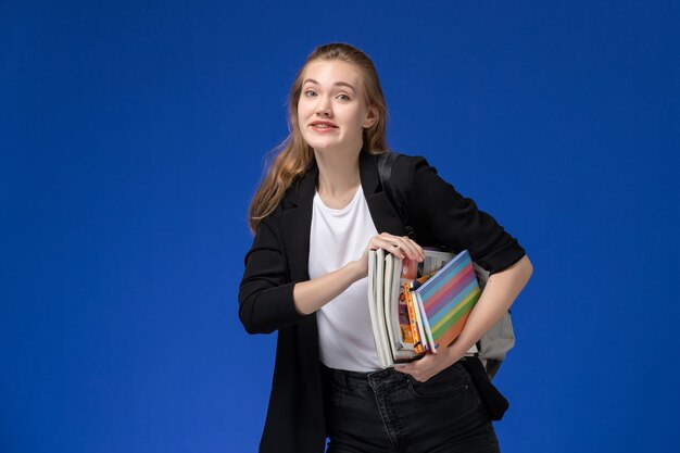 Studentessa di vista frontale in giacca nera che indossa uno zaino in possesso di libri e sorridente sul muro blu lezione scuola università college