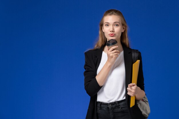 Studentessa di vista frontale in giacca nera che indossa zaino che tiene file giallo e caffè sulla lezione di università college scuola parete blu