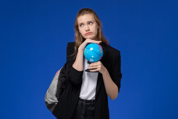 Studentessa di vista frontale in giacca nera che indossa zaino che tiene piccolo globo sulla lezione dell'università dell'università della scuola della parete blu