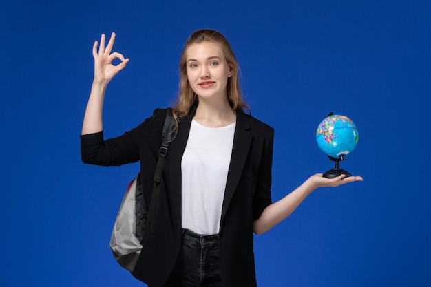 Studentessa di vista frontale in giacca nera che indossa zaino che tiene piccolo globo sulla terra blu chiaro dell'università dell'università della scuola della scuola della parete