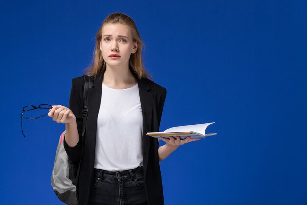 Studentessa di vista frontale in giacca nera che indossa zaino tenendo e leggendo il quaderno sulla parete blu lezioni scuola college università