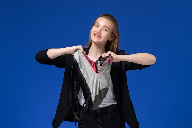 Studentessa di vista frontale in giacca nera che tiene zaino grigio sulle lezioni dell'università dell'università della scuola della parete blu