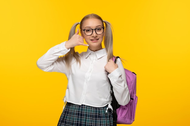 Studentessa felice bionda della giornata mondiale del libro in uniforme su sfondo giallo che mostra il segno del gesto di chiamata