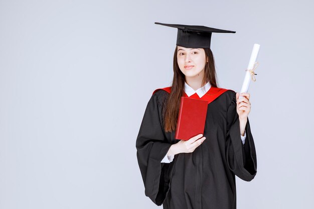 Studentessa in abito accademico che tiene libro e diploma. Foto di alta qualità