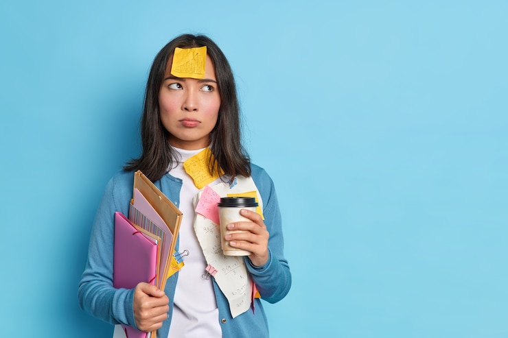 Foto gratuito studentessa intelligente ha un'espressione dispiaciuta seria scrive memo su adesivi di avviso coinvolti nel processo di apprendimento tiene una tazza di caffè usa e getta note informazioni sul progetto si trova sopra il muro blu