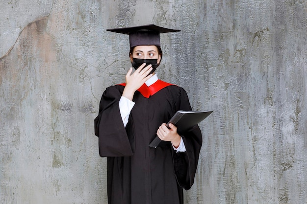 Studentessa laureata in maschera in piedi sopra il muro grigio. Foto di alta qualità