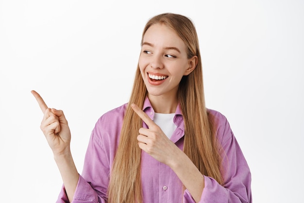 Studentessa moderna allegra che guarda e che punta il dito a sinistra sorridente mentre fissava la pubblicità sullo spazio della copia in piedi in abiti casual su sfondo bianco
