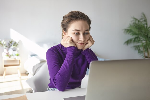 Studentessa seria concentrata che tiene le mani sotto le guance mentre impara online, ascolta lezioni su Internet o legge articoli scientifici