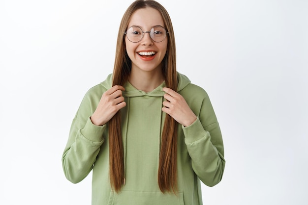 Studentessa sorridente con gli occhiali con i capelli lunghi che ride e sembra felice di provare nuovi occhiali in piedi in felpa con cappuccio su sfondo bianco