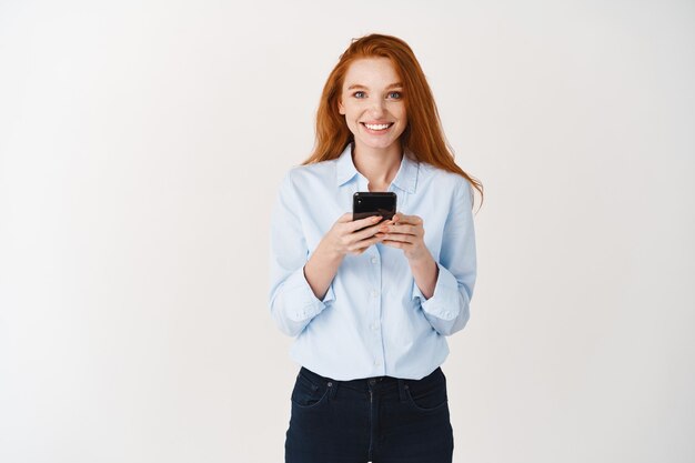 Studentessa sorridente con i capelli rossi e gli occhi azzurri usando il telefono cellulare, guardando felice davanti, in piedi sul muro bianco