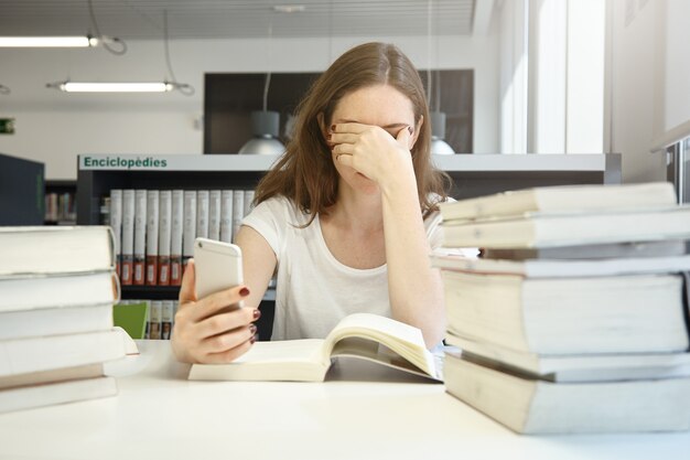 Studentessa stanca della scuola di economia seduta in biblioteca stropicciandosi gli occhi, annoiata di leggere il manuale di audit, controllando il tempo usando la sua applicazione per smartphone, circondata da pile di libri