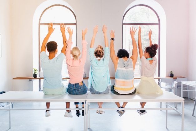 Studenti divertenti in posa con le mani in alto per l'album fotografico prima della laurea. Foto dell'interno dal retro di persone sedute davanti alla grande finestra in appartamento luminoso.