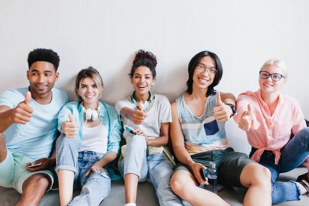 Studenti internazionali seduti sul pavimento e in posa con il pollice in su. Felici amici universitari in abiti eleganti che si divertono nel loro campus dopo le lezioni ..