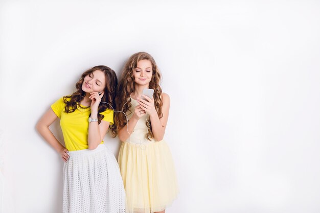 Studio fotografico di due ragazze sorridenti in piedi che ascoltano musica su uno smartphone e si divertono. La bruna indossa una gonna bianca e una maglietta gialla, e la ragazza bionda indossa un vestito giallo.