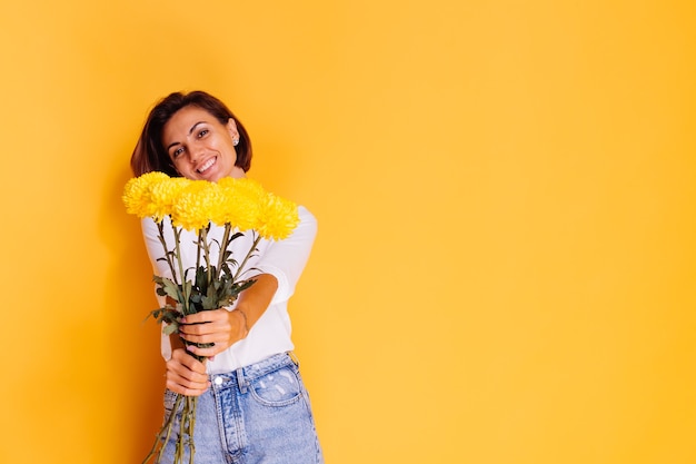 Studio girato su sfondo giallo Felice indoeuropea donna capelli corti indossando abiti casual, camicia bianca e pantaloni in denim azienda bouquet di astri gialli