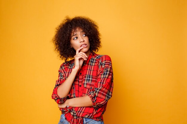 Studio immagine di pensare bella giovane donna africana in posa isolato su sfondo arancione vestito in camicia rossa.