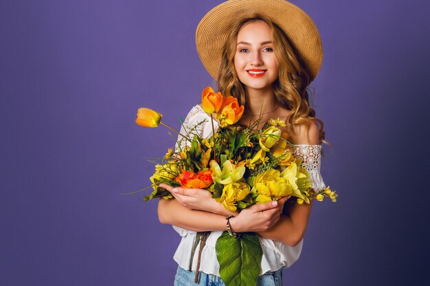 Studio moda ritratto di donna bionda piuttosto carina in cappello di paglia, camicia di cotone bianco seduto e in possesso di bouquet di fiori primaverili incredibili. Indossa un elegante abito retrò.