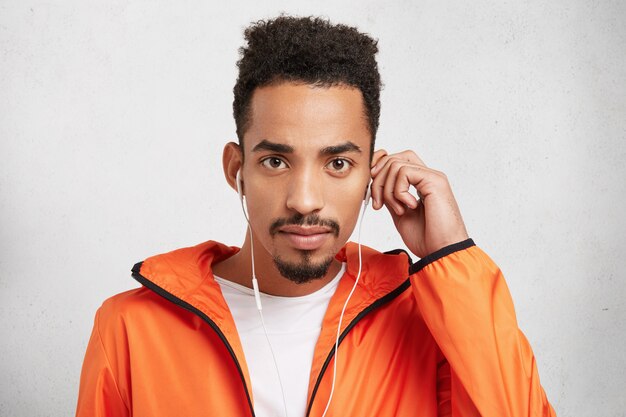 Studio shot di Afro American uomo con elegante pettinatura, indossa arancione giacca a vento alla moda, ascolta le canzoni in auricolari,