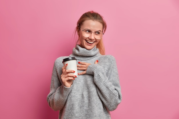 Studio shot di allegra ragazza europea ridacchia positivamente mantiene la mano sul petto e tiene il caffè da asporto