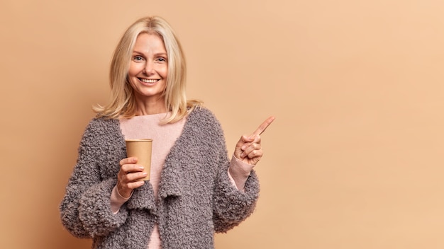 Studio shot di bella bionda cinquantenne donna sorride positivamente tiene la carta usa e getta tazza di bevanda calda indossa pelliccia indica lontano isolato su muro marrone