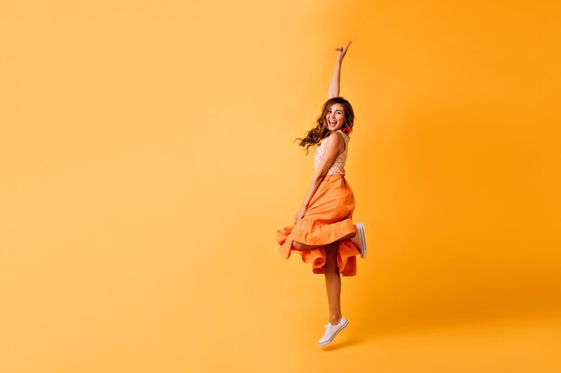 Studio shot di bella ragazza in gonna arancione e scarpe bianche. Eccitata signora dai capelli rossi che salta sul giallo.
