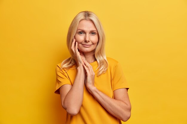 Studio shot di calma bella donna con pelle sana tocca il viso indossa delicatamente il trucco minimo ha un tenero sorriso si prende cura della sua carnagione indossa maglietta gialla in un tono con lo sfondo.