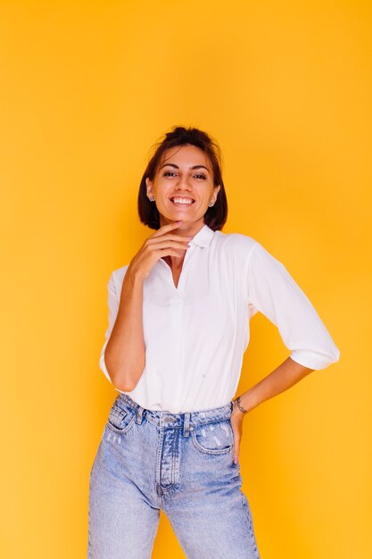 Studio shot di donna felice capelli corti indossando una camicia bianca e pantaloni in denim in posa sulla parete gialla