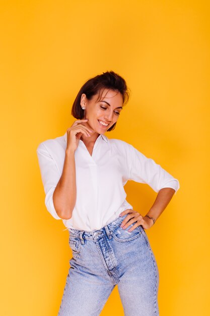 Studio shot di donna felice capelli corti indossando una camicia bianca e pantaloni in denim in posa sulla parete gialla