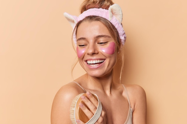 Studio shot di donna felice lava il corpo con sorrisi di spazzola da bagno posa volentieri a torso nudo indossa fascia applica cerotti idrogel sotto gli occhi isolati su sfondo beige. Concetto di cura della pelle del viso