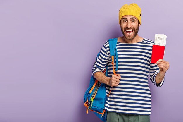 Studio shot di felice con la barba lunga maschio backpacker detiene il passaporto con documento, trasporta lo zaino sulla spalla, sorride volentieri, vestito con un abbigliamento casual, isolato sopra la parete viola, pronto per il viaggio