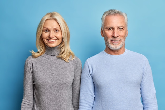 Studio shot di felice donna matura e uomo esprime emozioni positive godersi la vita aspettare che i bambini vengano vestiti in poloneck casual isolato sopra il muro blu dello studio