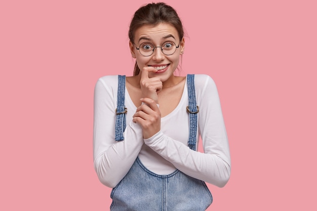 Studio shot di felice giovane donna tiene le mani vicino ai denti, sorride ampiamente, sente notizie positive dall'interlocutore