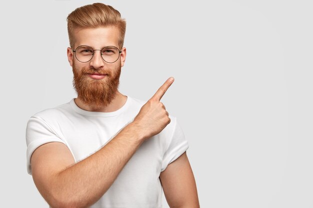 Studio shot di ginger hipster con folta barba, taglio di capelli alla moda, ha un'espressione seria, punti con il dito indice nell'angolo in alto a destra