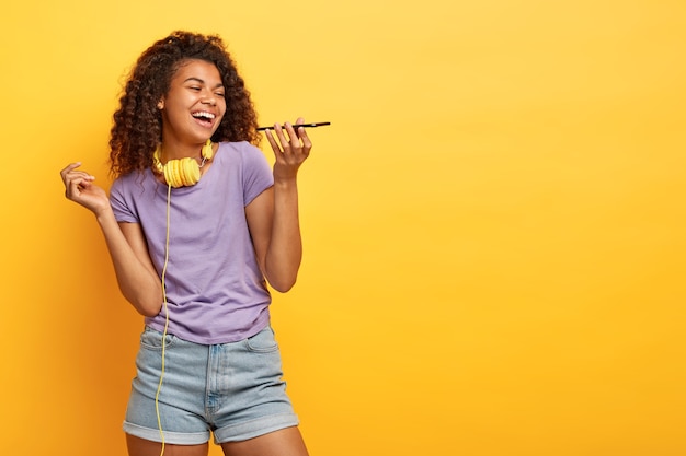 Studio shot di gioiosa giovane femmina con acconciatura afro in posa contro il muro giallo