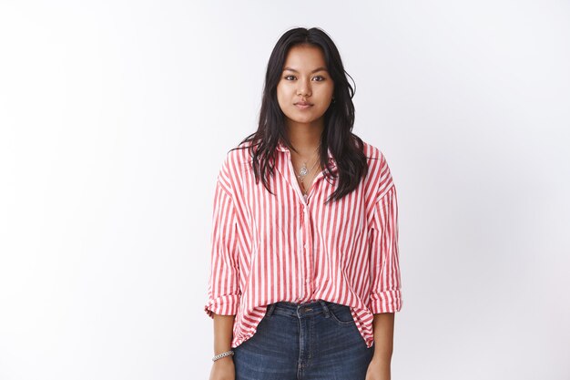 Studio shot di giovane donna vietnamita in camicetta a righe rosa e jeans in piedi rilassato mentre fissava la telecamera focalizzata, tenendo le braccia al lato con espressione casual in posa contro il muro bianco