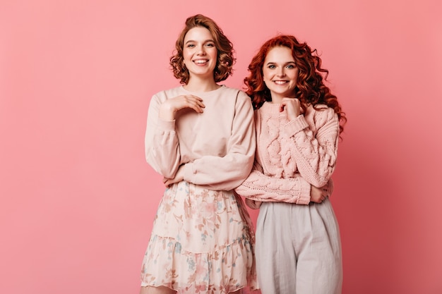 Studio shot di incantevoli ragazze isolate su sfondo rosa. Due amici alla moda che esaminano macchina fotografica con il sorriso.