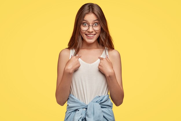 Studio shot di indignato giovane bruna con gli occhiali in posa contro il muro giallo