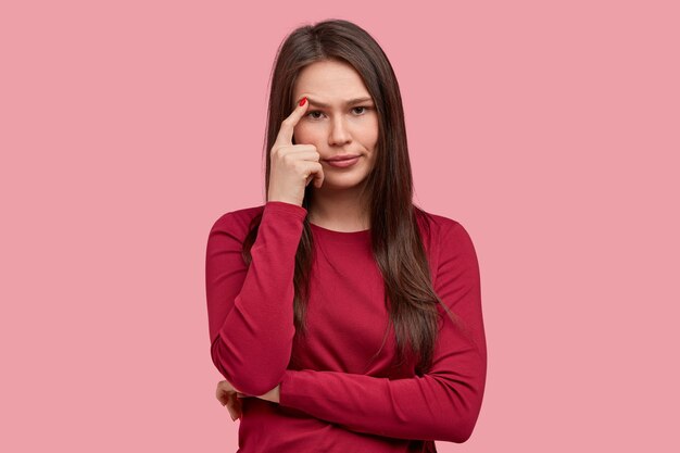 Studio shot di pensieroso giovane donna mantiene il dito anteriore vicino alle sopracciglia, indossa un maglione rosso, tiene le mani incrociate