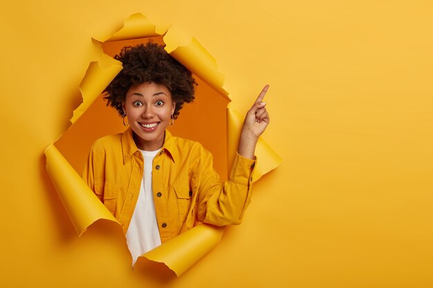 Studio shot di positivo donna afro-americana punta il dito per copiare lo spazio sopra, eccitato da una buona informazione, sorride piacevolmente, indossa una giacca gialla, si trova nel buco della carta strappata.