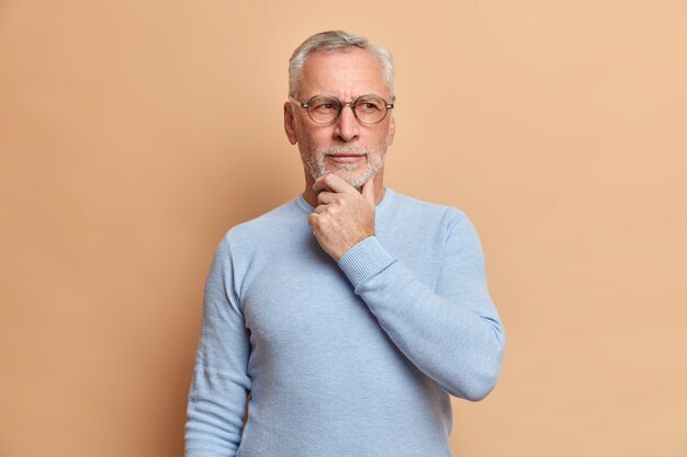 Studio shot di premuroso uomo senior tiene il mento contempla qualcosa di vestito casualmente cerca di ricordare qualcosa e raccogliere con pensieri pone contro il muro marrone