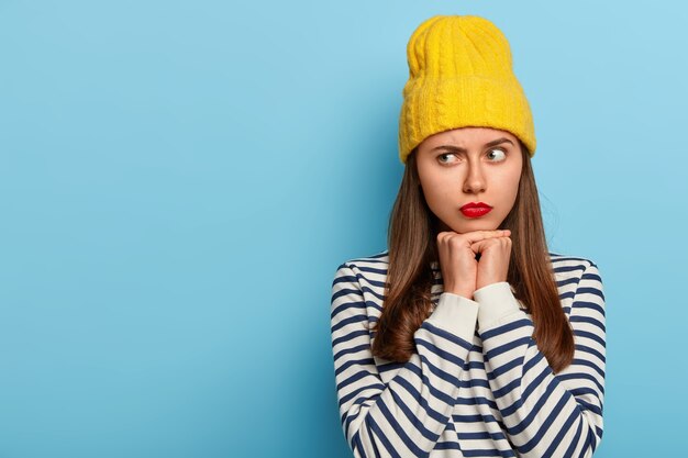 Studio shot di riflessivo giovane modello femminile guarda da parte con espressione seria, vestito con un maglione a strisce, giallo cappello elegante, isolato su sfondo blu, uno spazio vuoto per promo