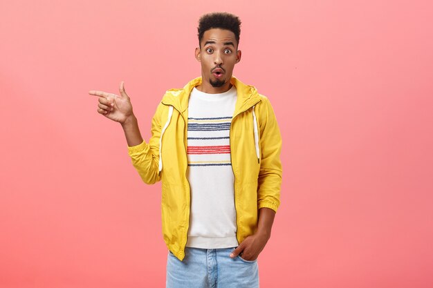 Studio shot di sorpreso impressionato elegante afroamericano studente maschio in giacca gialla alla moda piegando le labbra nel suono wow alzando le sopracciglia dallo stupore che punta a sinistra interrogato sul muro rosa