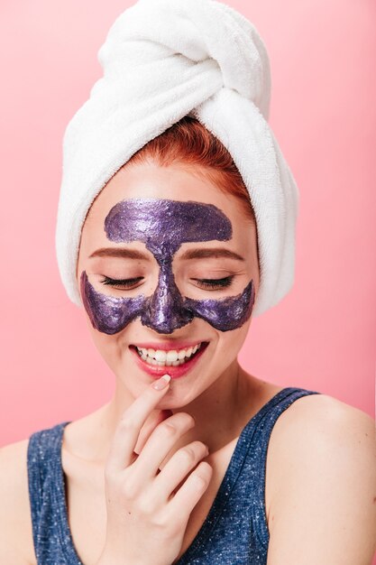 Studio shot di spensierata ragazza che ride durante il trattamento termale. Modello femminile emotivo con maschera facciale in posa su sfondo rosa.