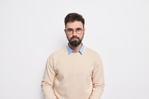 Studio shot di un bell'uomo europeo adulto barbuto guarda direttamente, con un'espressione seria ha un viso determinato vestito con occhiali rotondi con maglione pulito