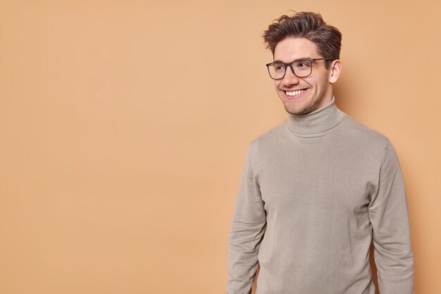 Studio shot di un bell'uomo europeo sorride positivamente essendo di buon umore concentrato da qualche parte indossa occhiali e posa a collo alto su sfondo beige con spazio per la copia per la tua promozione