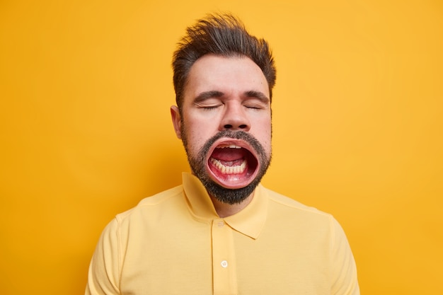 Studio shot di un uomo caucasico stanco barbuto chiude gli occhi tiene la bocca ampiamente aperta mentre lo sbadiglio ha un'espressione assonnata vestita di camicia