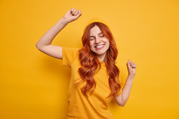 Studio shot di una donna rossa spensierata alza le danze a mano e sorride allegramente si diverte indossando un cappello e una maglietta casual