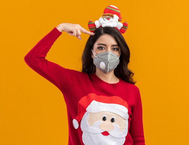 Stufa giovane ragazza carina che indossa la fascia di Babbo Natale e un maglione con maschera protettiva che fa gesto di suicidio isolato sul muro arancione